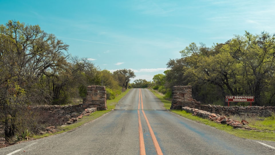 A road leading to Lake Brownwood State Park with stone walls and trees on either side.