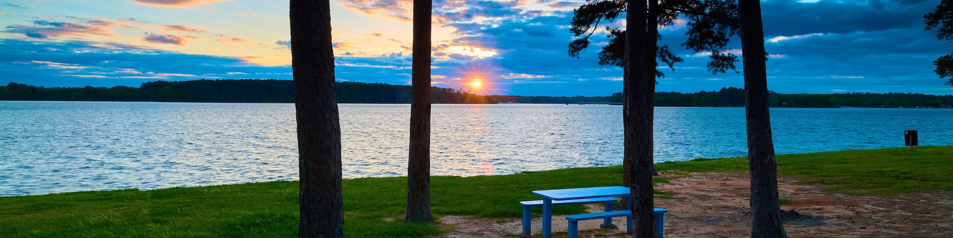 Escena junto a un lago con atardecer, árboles y una mesa para picnic.