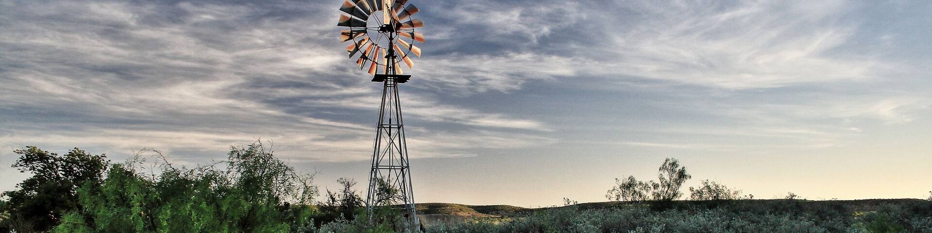 A windmill in a field with sparse vegetation under a cloudy sky.