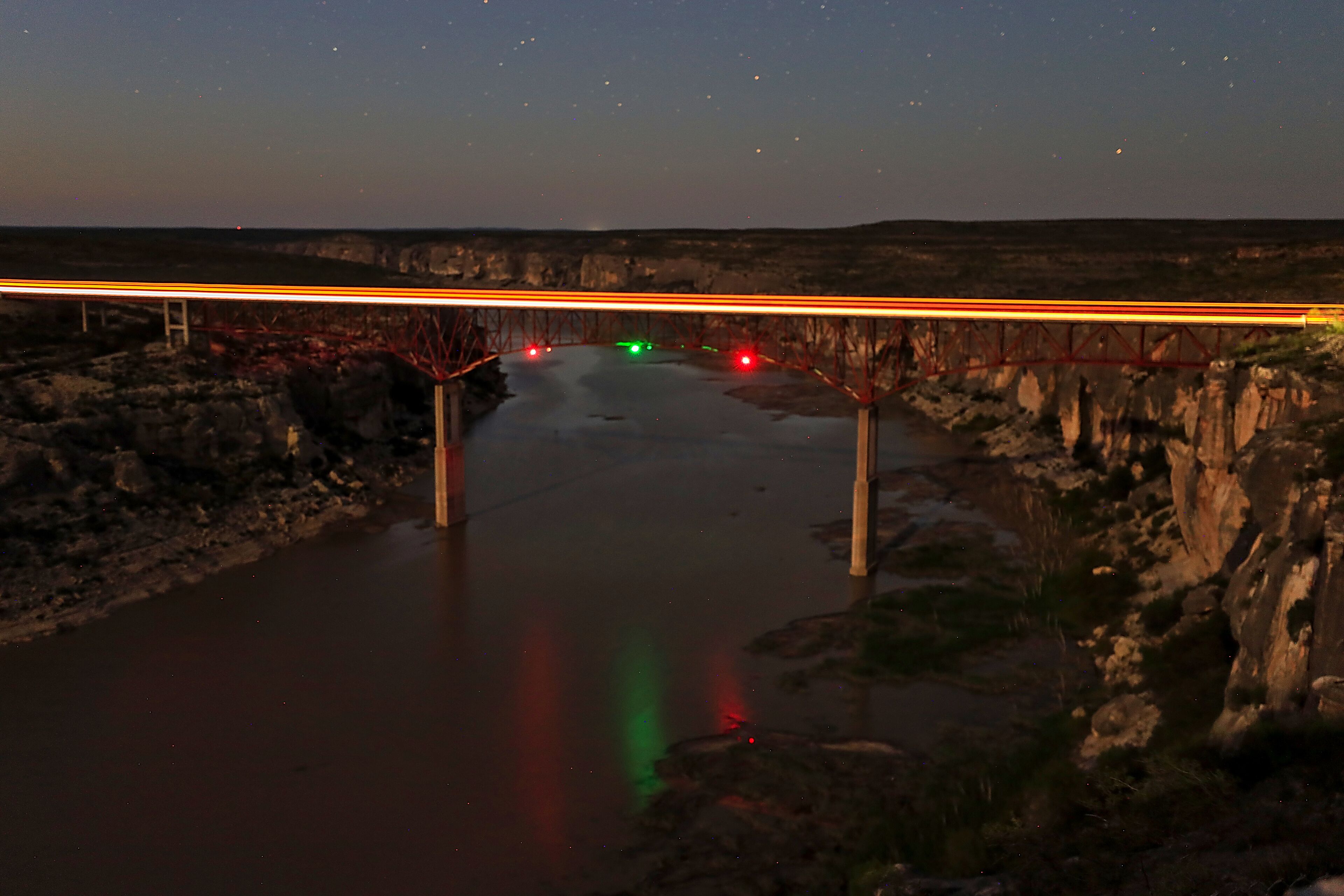 A bridge over a river at night with starry sky and reflections of lights on the water.