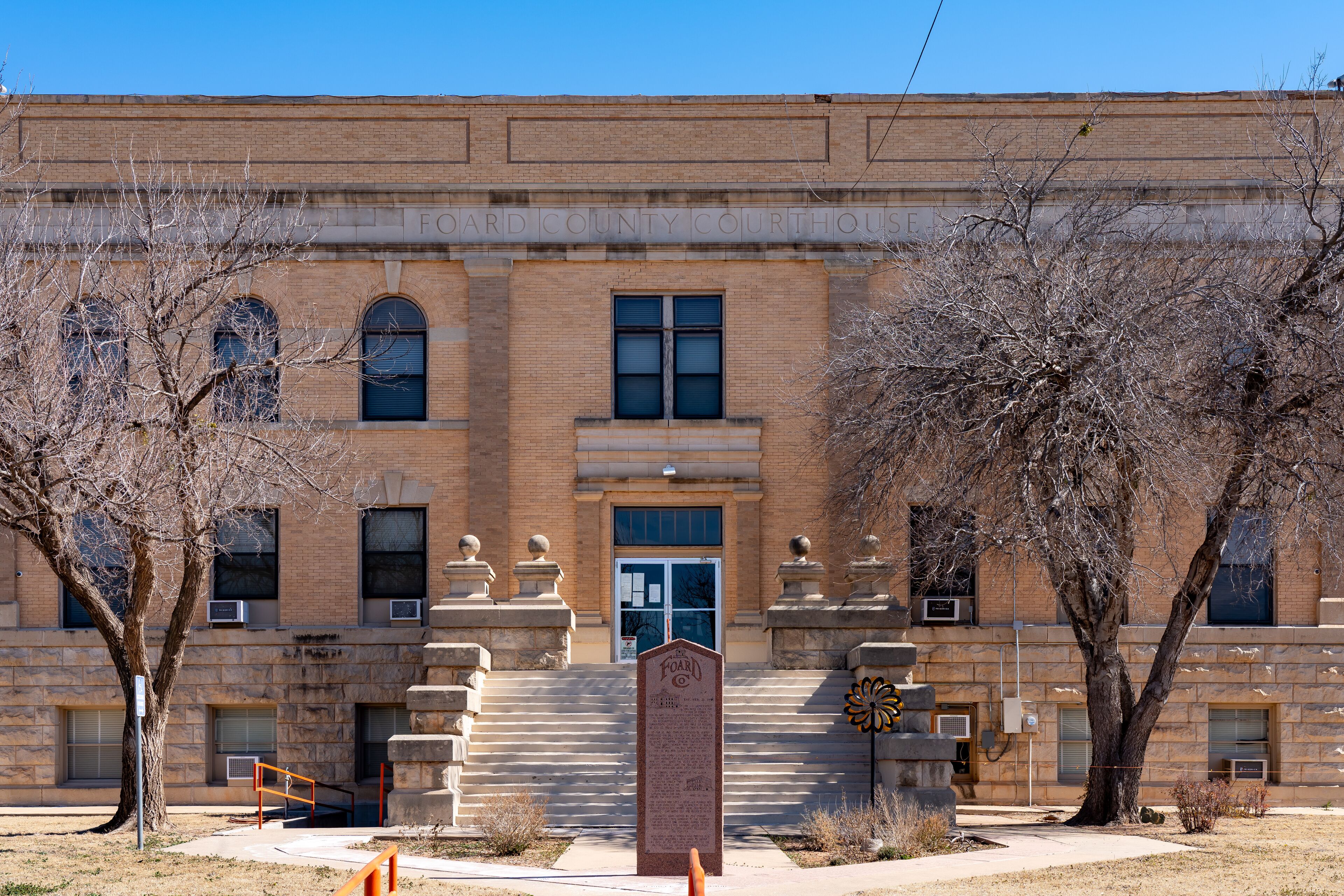 A brick courthouse with a central entrance, flanked by two large trees and a stone monument in front.