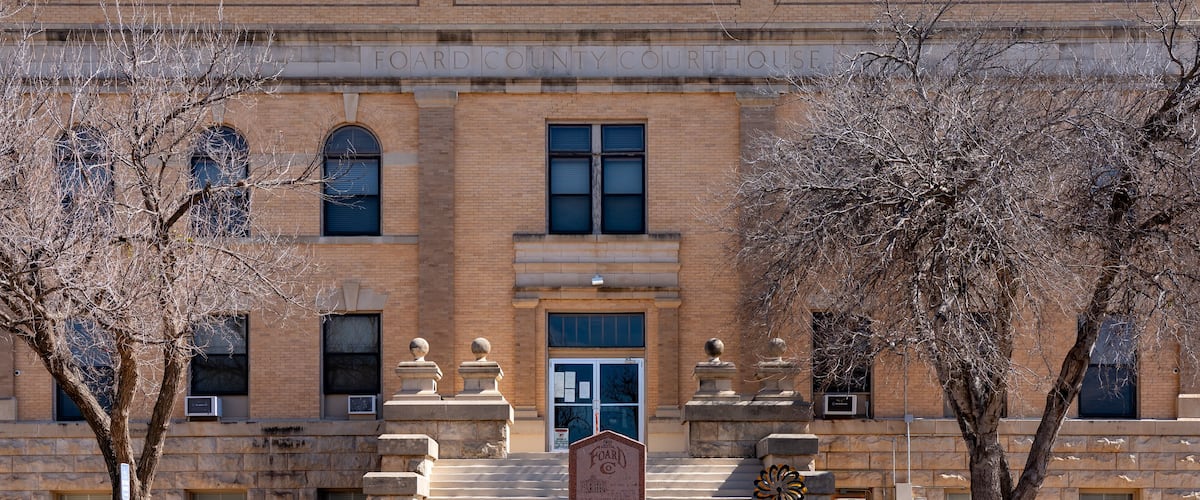 A brick courthouse with a central entrance, flanked by two large trees and a stone monument in front.