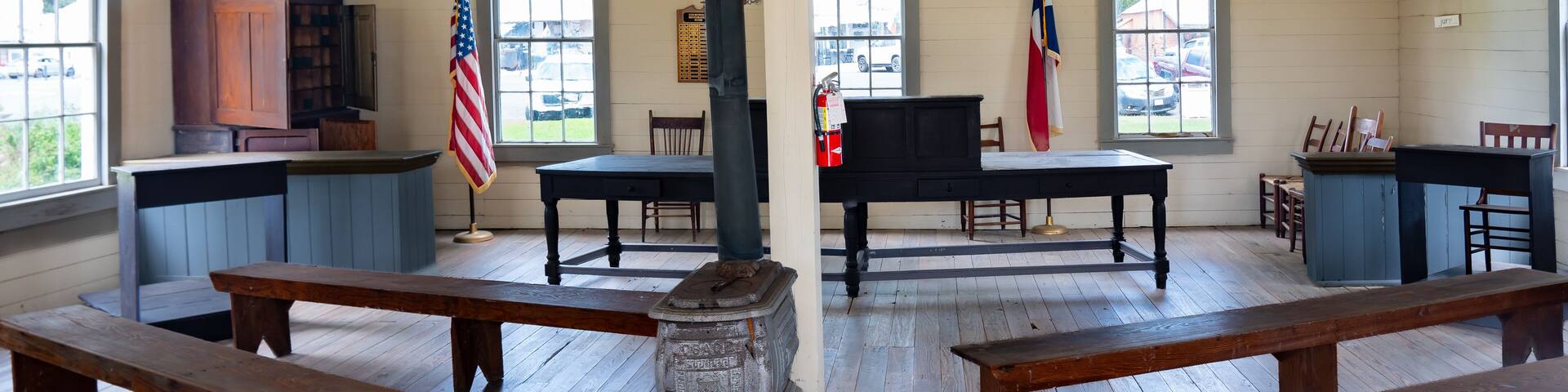 A historical room with wooden benches, a stove, a flag, and a piano.