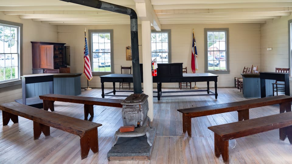 A historical room with wooden benches, a stove, a flag, and a piano.