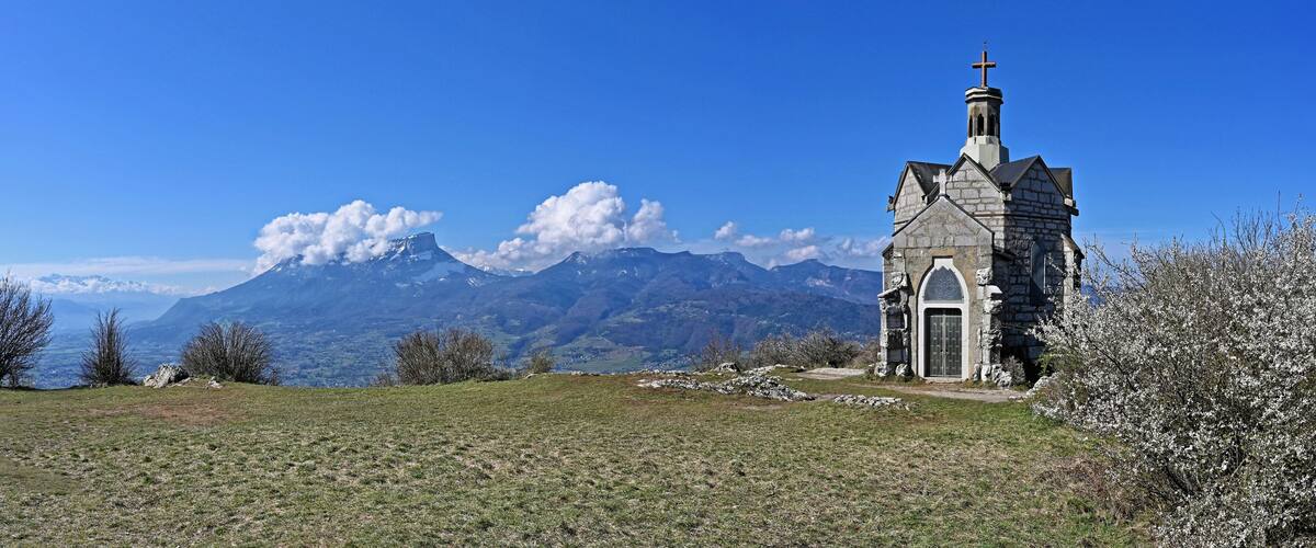 A stone church with a cross on top, surrounded by a grassy field and mountains in the background.