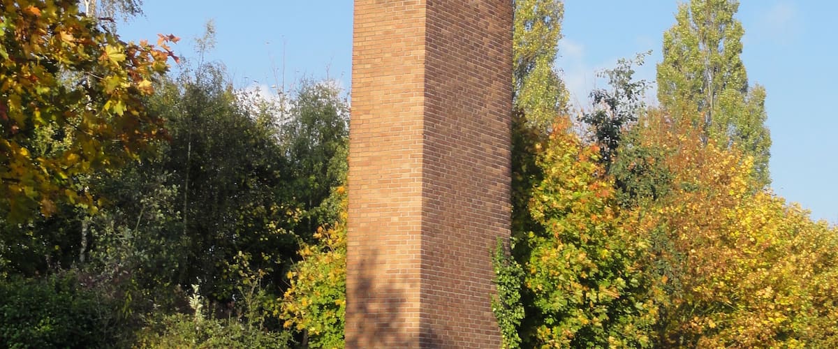 A tall brick chimney with a metal plaque, surrounded by trees with autumn leaves and a metal fence.