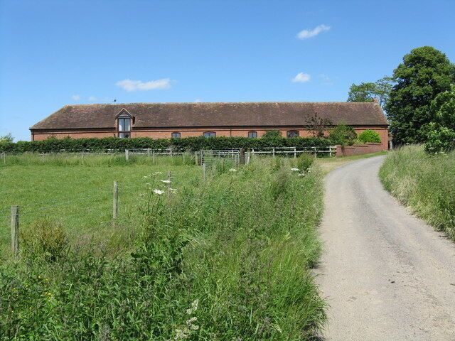 A rural road leading to a red brick farmhouse with a thatched roof, surrounded by green fields and a fence.