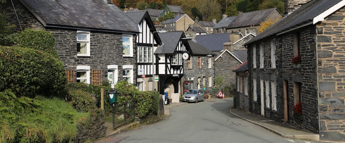 A quaint village street lined with stone and timber-framed houses, a car parked on the side, and a backdrop of autumn-colored trees.
