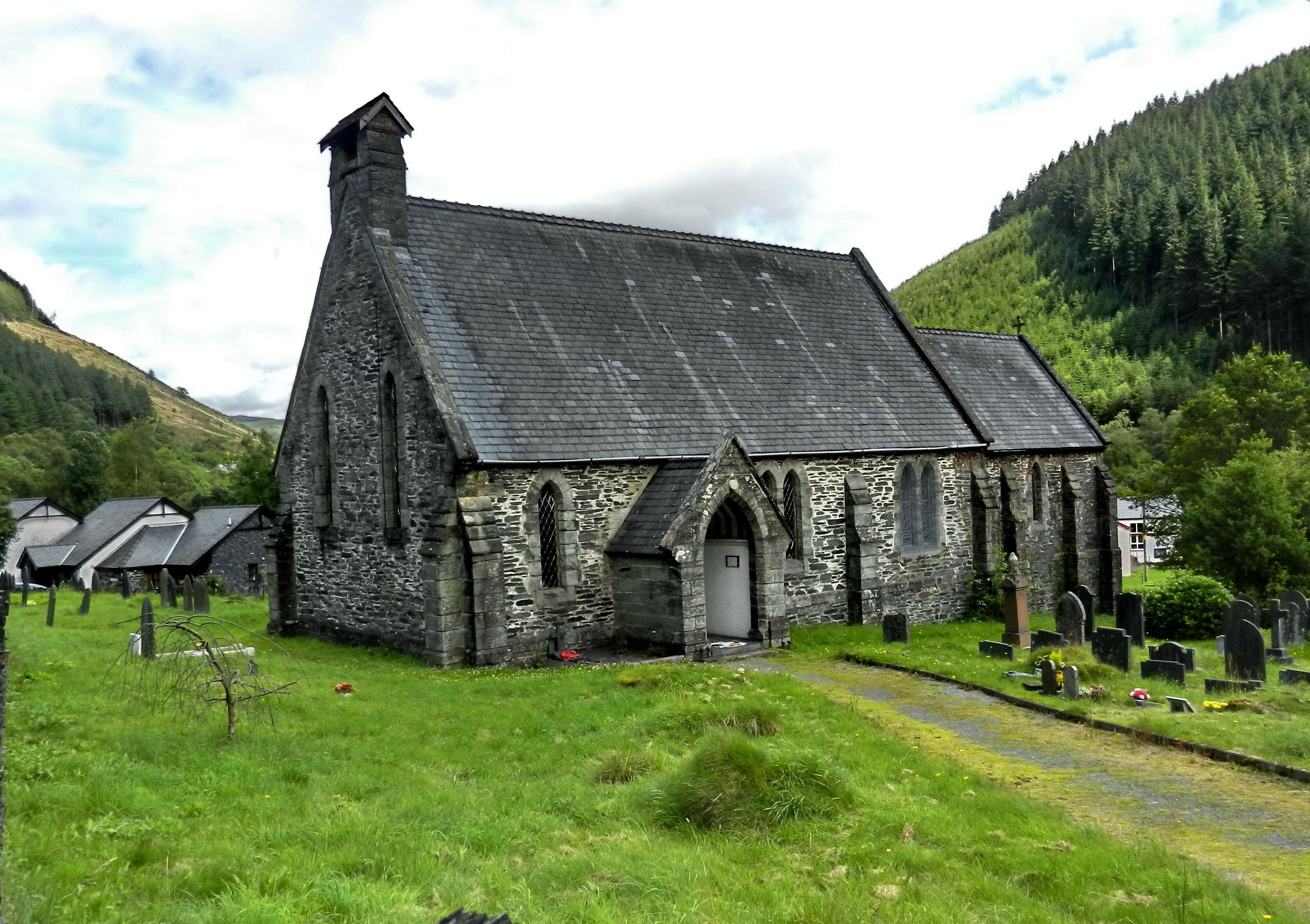 A stone church with a steep roof and arched windows, surrounded by a cemetery and a forested hillside.