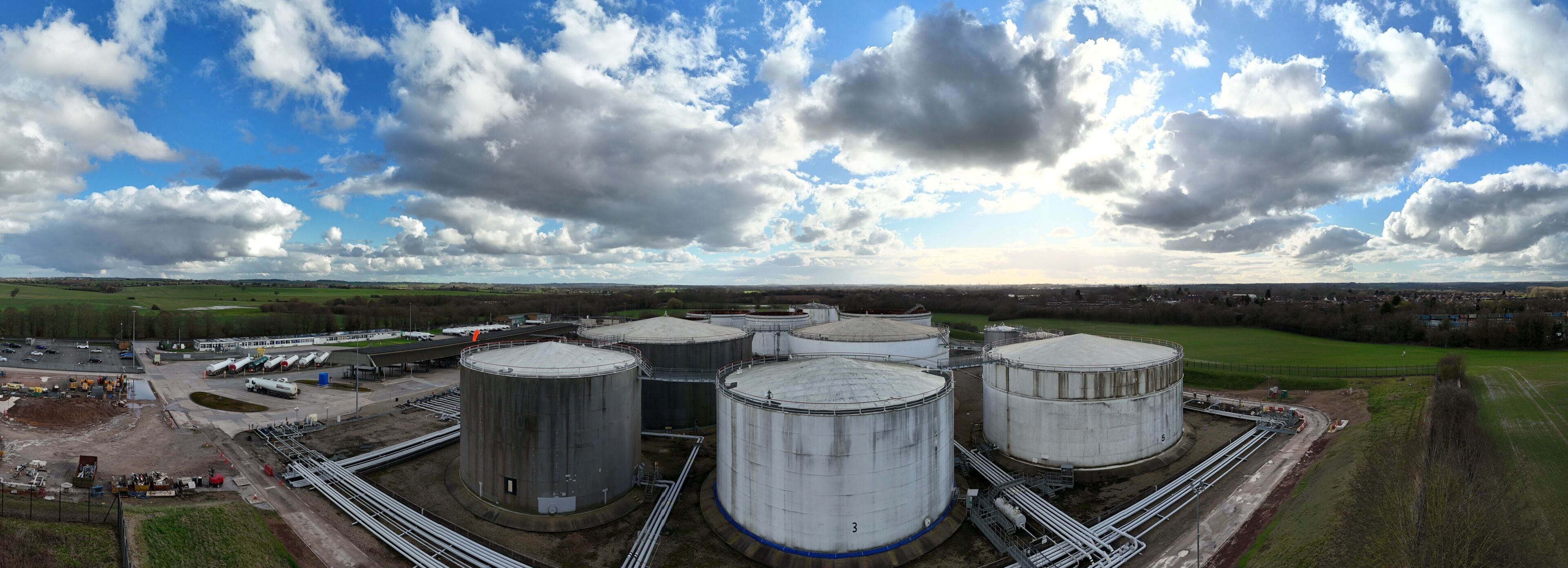 Aerial view of cylindrical oil tanks gleaming under the expansive sky contrast against the lush green fields, Kingsbury Oil Terminal, Tamworth, United Kingdom.