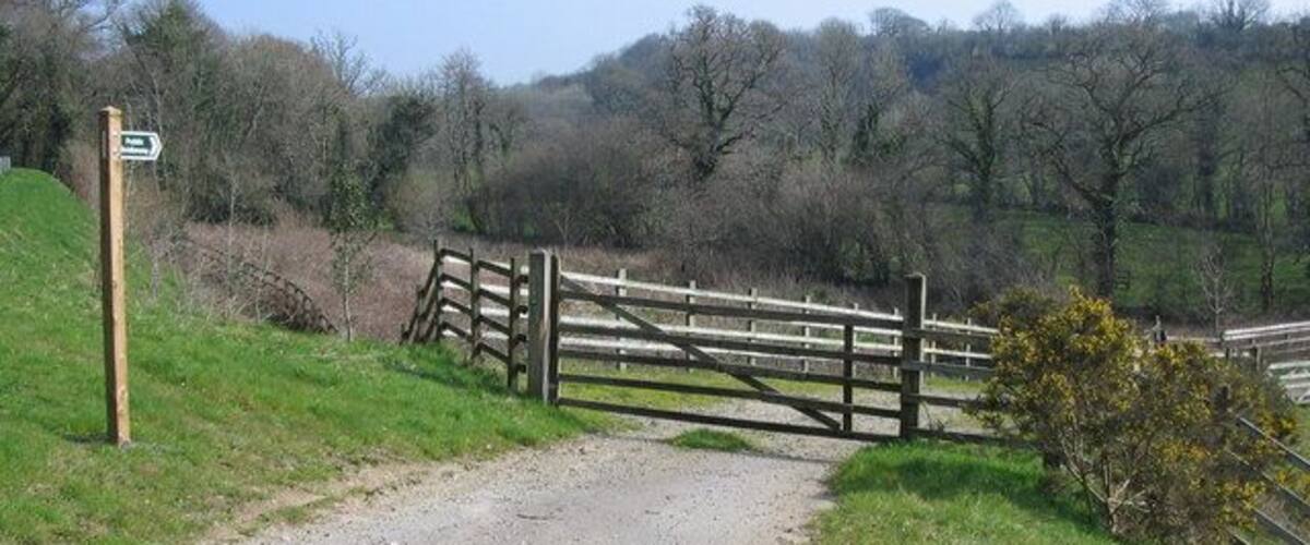 A gravel path leading to a wooden gate in a rural setting with trees and hills in the background.
