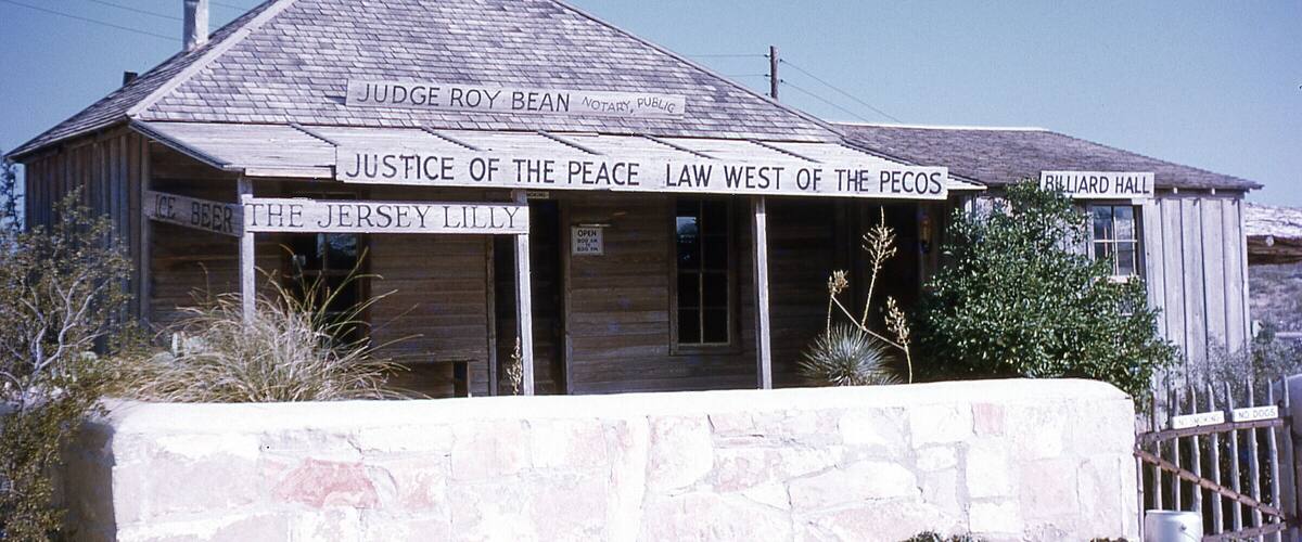 A wooden building with signs reading "Judge Roy Bean Notary Public," "Justice of the Peace Law West of the Pecos," and "Billiard Hall."