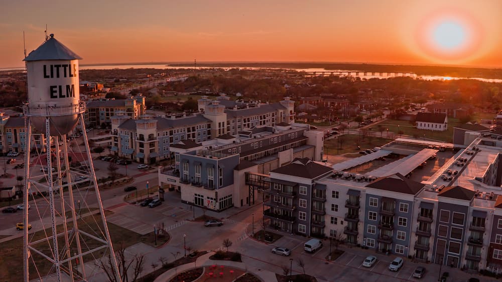 Un château d’eau portant l’inscription « LITTLE ELM » et un quartier résidentiel avec des immeubles et un parking, le tout baigné par les couleurs du coucher de soleil.