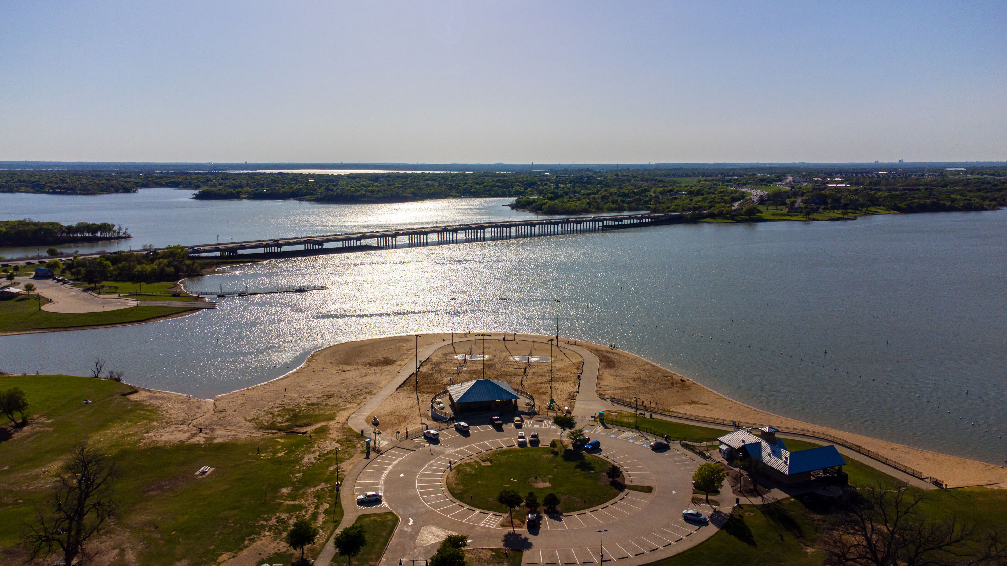 Aerial view of a sun-kissed beach meeting the vast lake under a clear sky, with a bridge stretching across the water, Little Elm, Texas, United States.