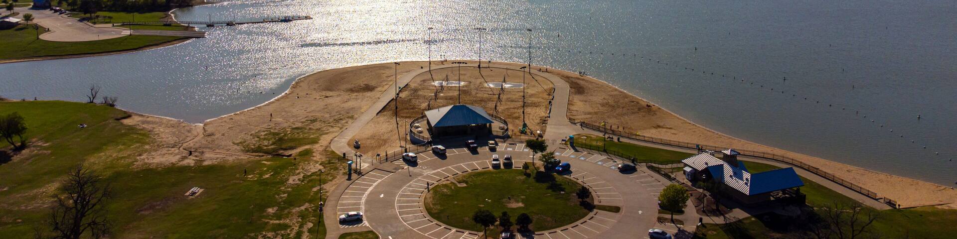 Aerial view of a sun-kissed beach meeting the vast lake under a clear sky, with a bridge stretching across the water, Little Elm, Texas, United States.