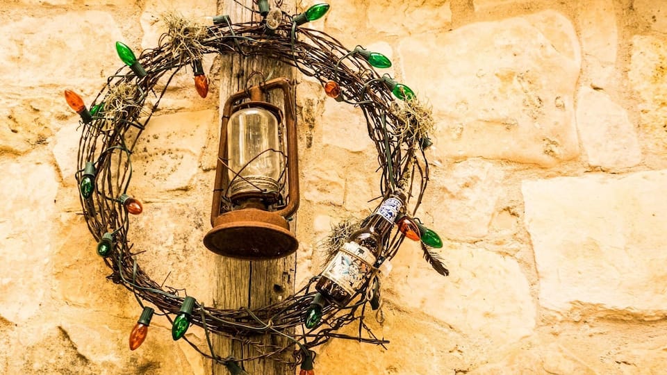 A wreath with Christmas lights and a lantern on a stone wall.