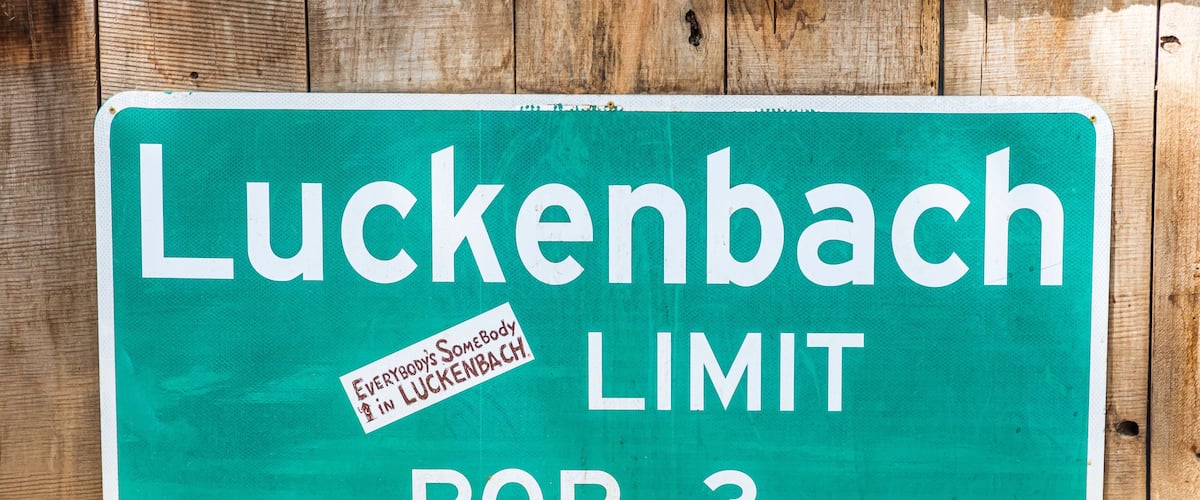 A green sign with white text that reads "Luckenbach LIMIT POP. 3" on a wooden background.