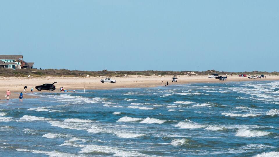 A beach with waves, people, and houses in the background.