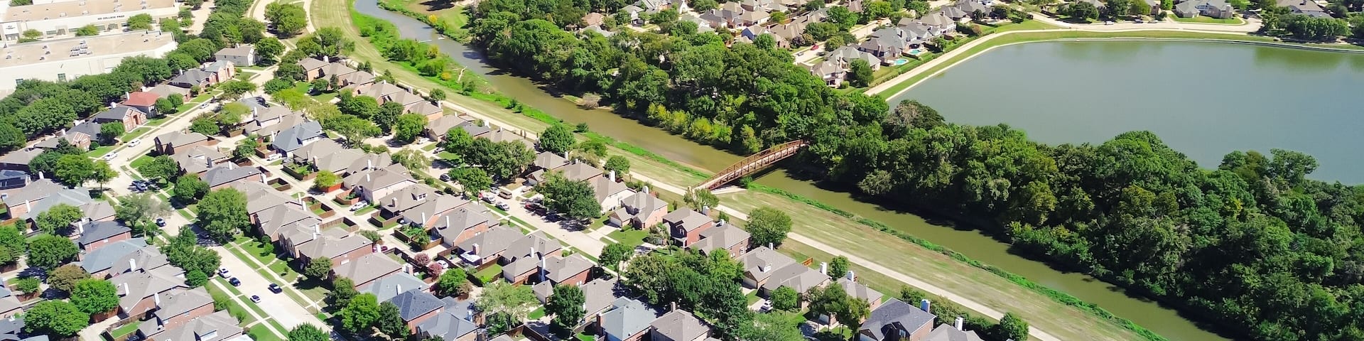 Coppell Texas suburban park lush green trees, lake, creek near upscale residential neighborhood row of two-story houses with pools, interexchange stack and downtown Dallas in background, aerial