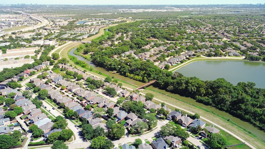 Coppell Texas suburban park lush green trees, lake, creek near upscale residential neighborhood row of two-story houses with pools, interexchange stack and downtown Dallas in background, aerial