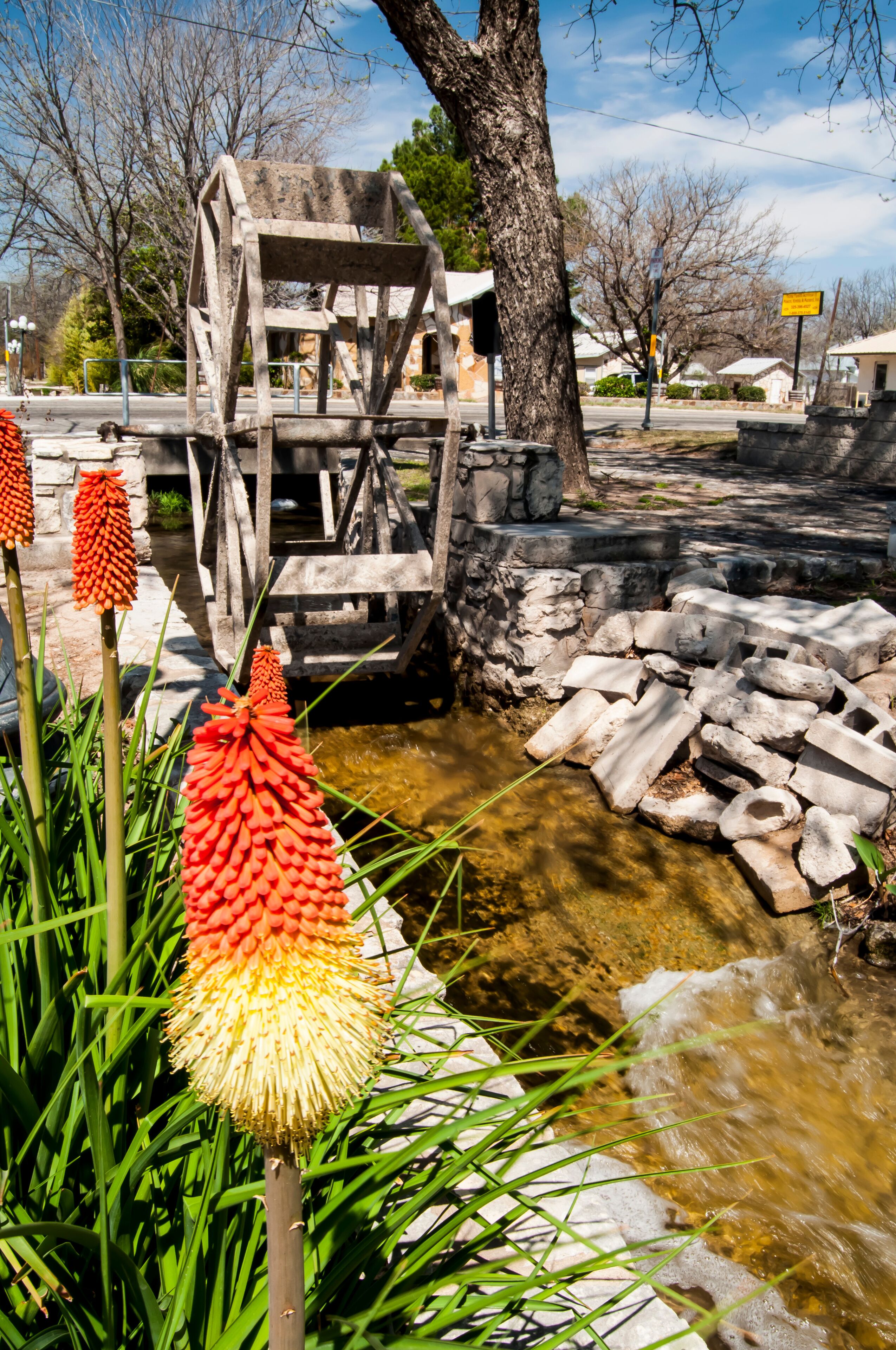 A wooden water wheel, a stone wall, and red flowers in a park.