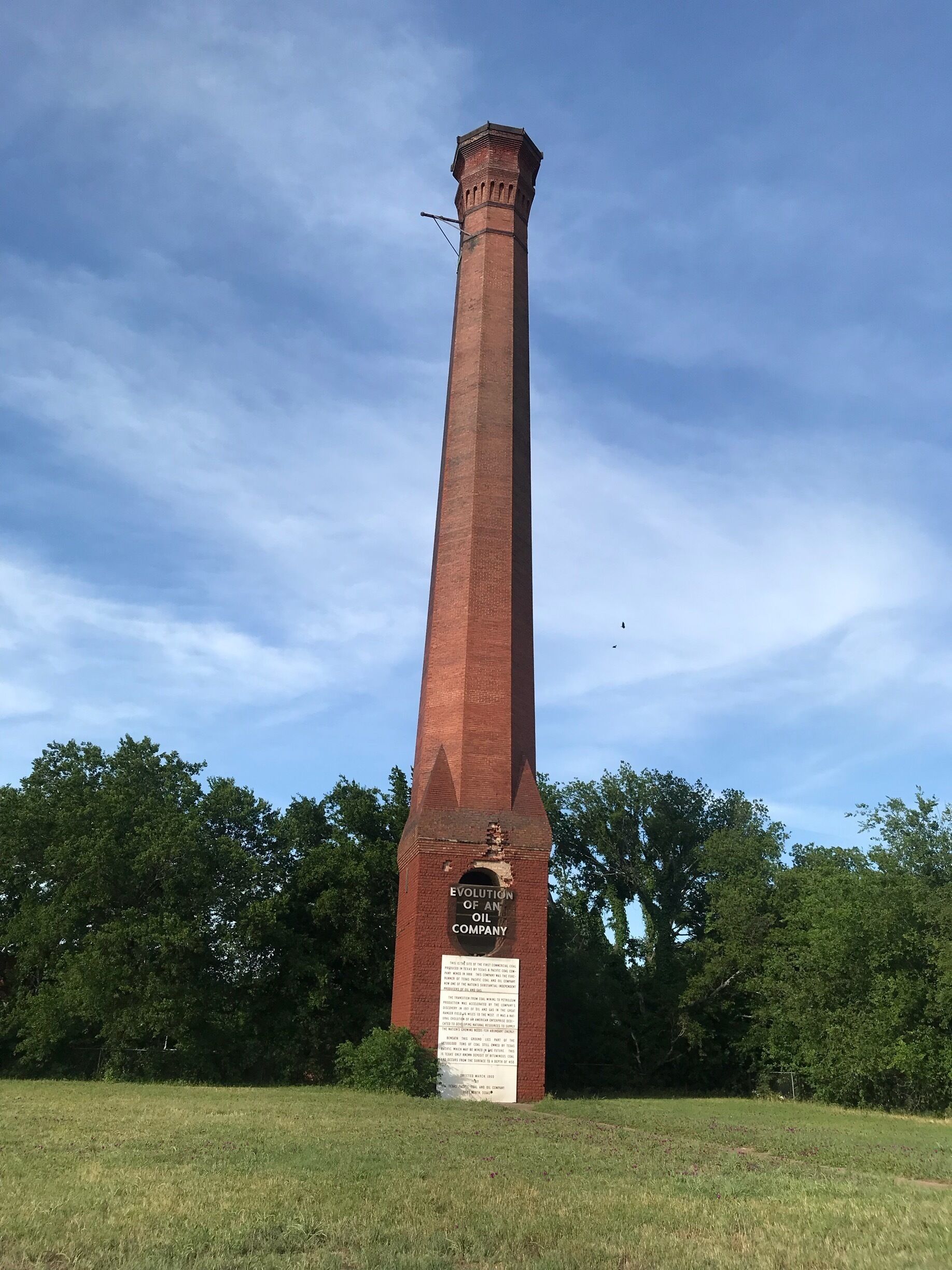 A tall brick monument with a plaque titled "Evolution of an Oil Company" in a grassy field with trees in the background.