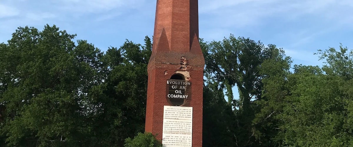 A tall brick monument with a plaque titled "Evolution of an Oil Company" in a grassy field with trees in the background.
