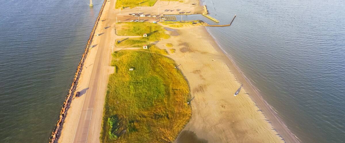 An aerial view of a coastal area with a sandy beach, grassy patches, and a road running parallel to the shore.