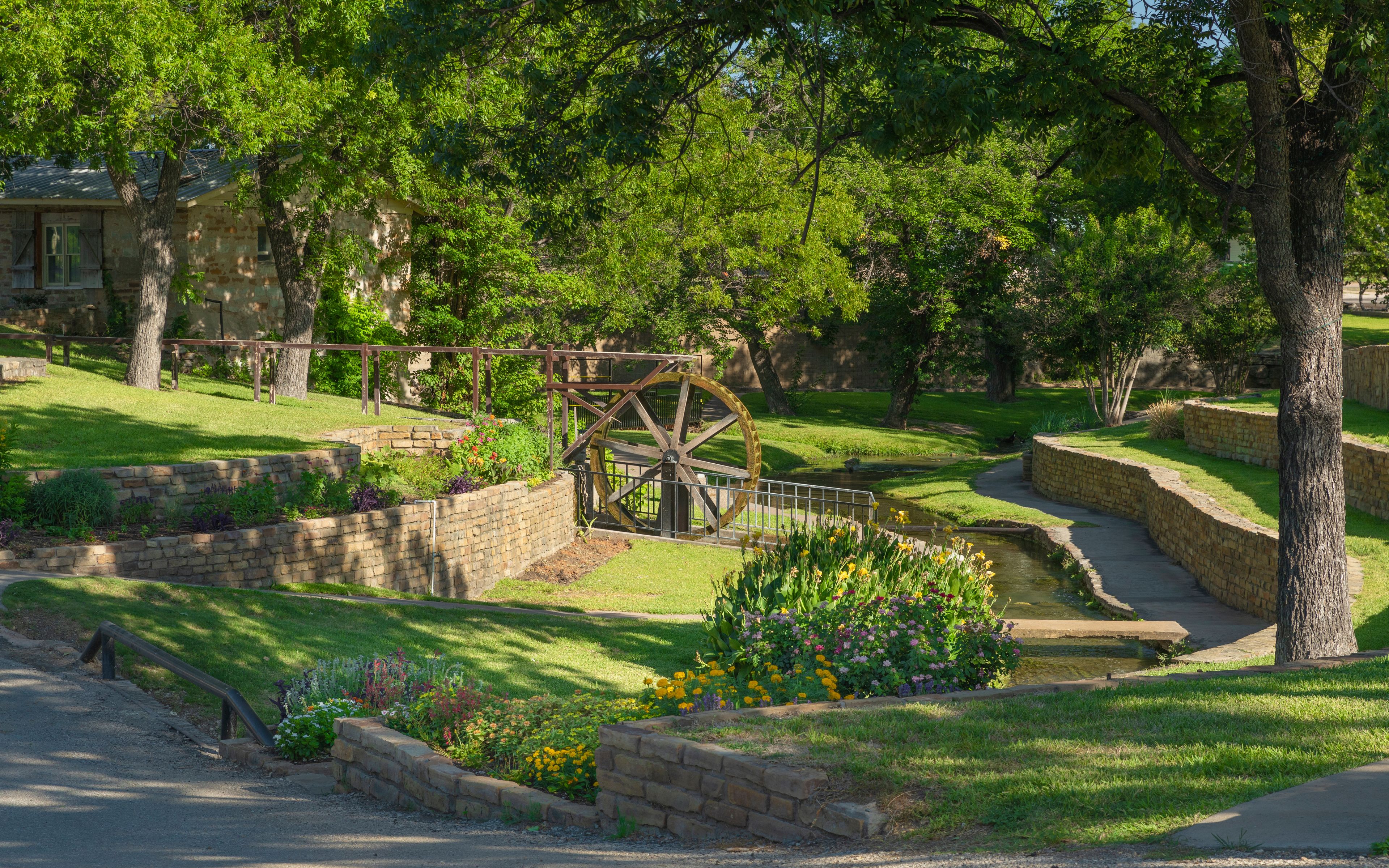 A historic water wheel, lush greenery, and stone walls in a park setting.