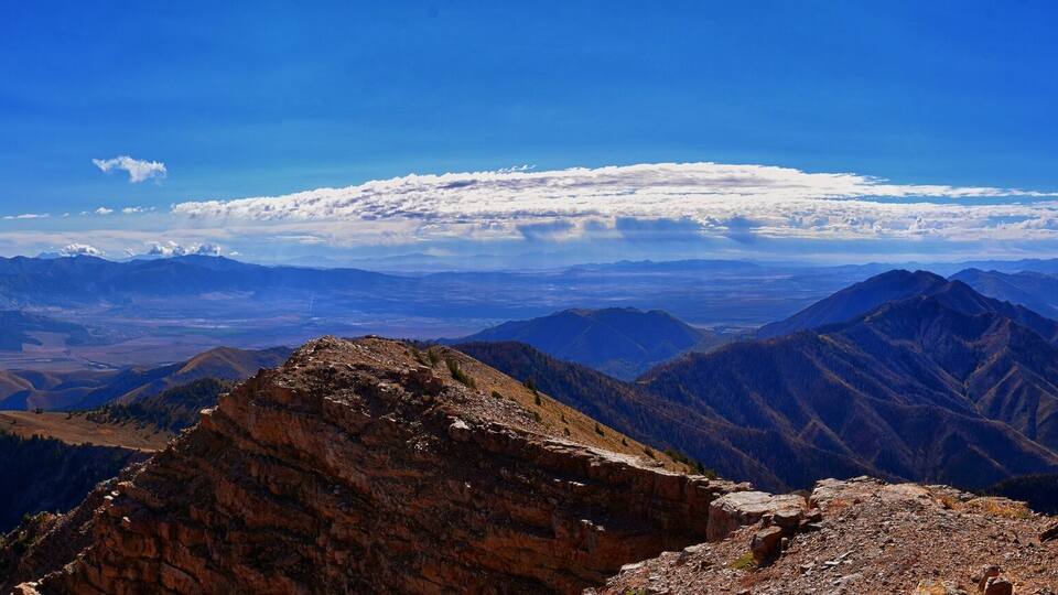 A mountain range with a snow-capped peak in the distance, rocky terrain in the foreground, and a clear blue sky.