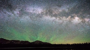 Céu estrelado sobre um lago tranquilo, com o contorno de uma montanha e uma floresta ao primeiro plano.