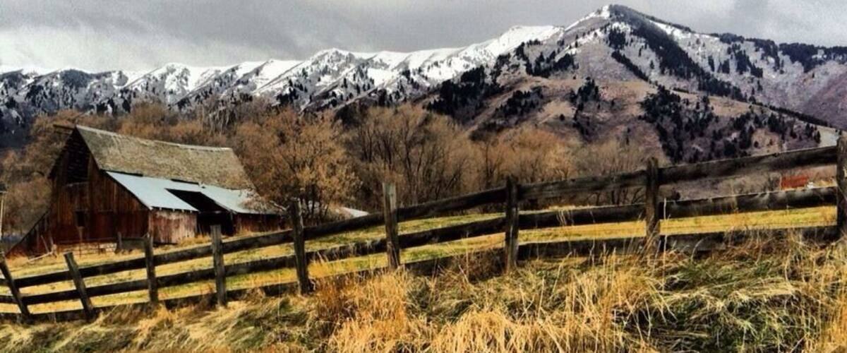 A rural landscape with a wooden fence, a barn, and snow-capped mountains in the background.