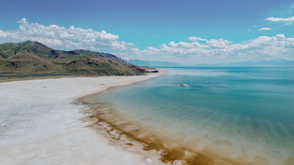 A coastal landscape with a sandy beach, clear turquoise waters, and distant mountains under a blue sky with scattered clouds.
