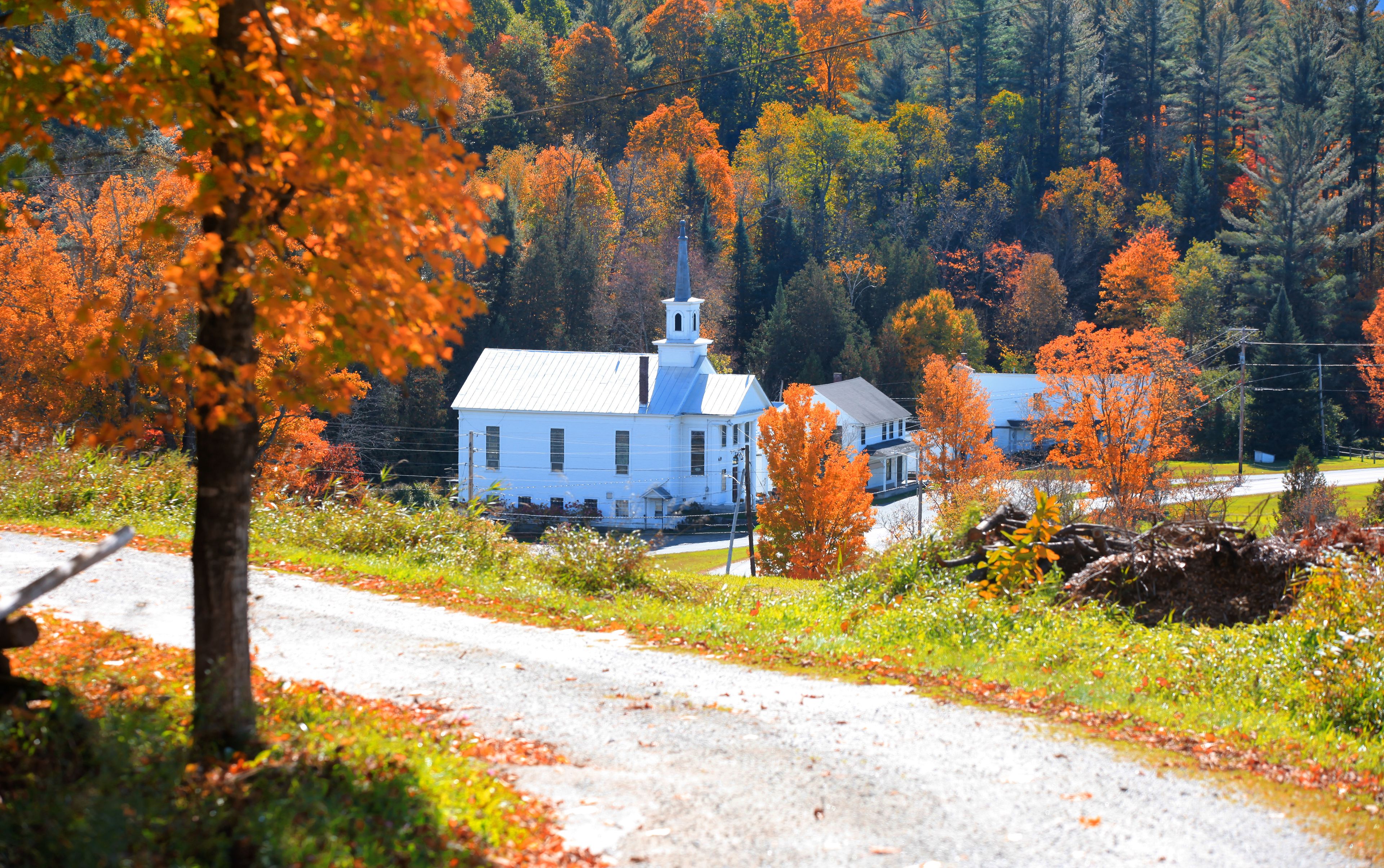 A white church with a steeple, surrounded by autumn foliage and a gravel road.