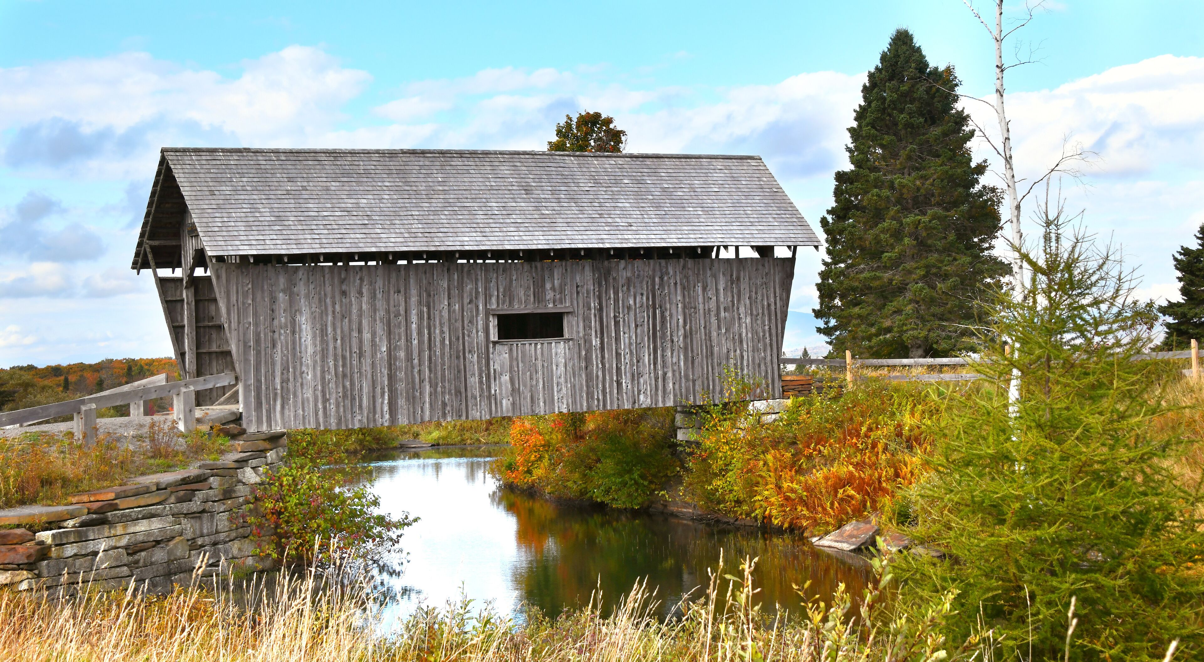 A wooden barn with a sloped roof, a small window, and a stone wall in the foreground.