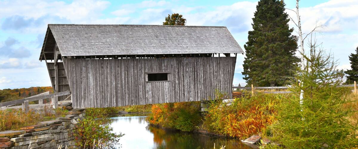 A wooden barn with a sloped roof, a small window, and a stone wall in the foreground.
