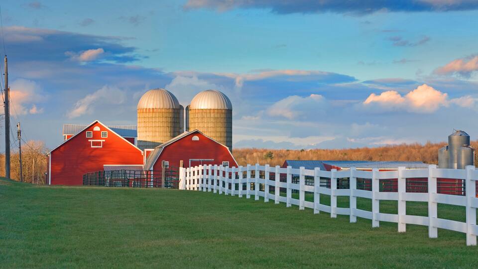 A red barn, two silos, and a white fence in a rural setting.