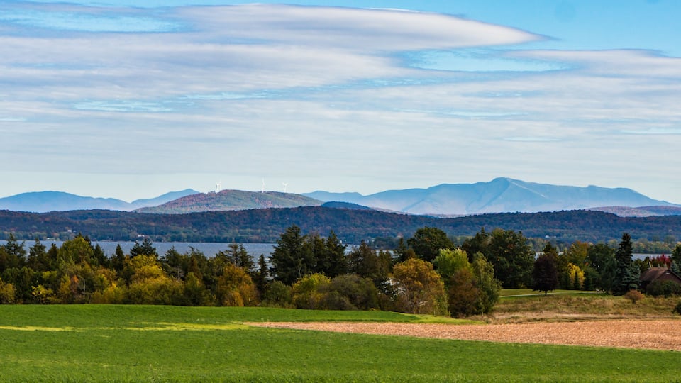 A scenic landscape with a lake, green fields, and distant mountains under a partly cloudy sky.