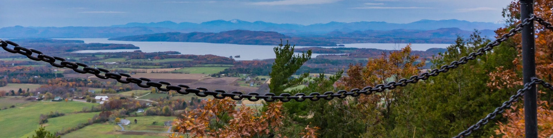Una cerca de malla metálica con vista a un valle cubierto de follaje otoñal, un lago y montañas distantes.