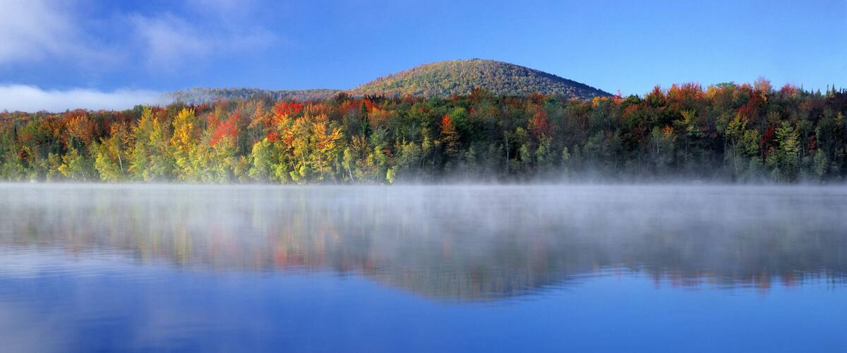 A calm lake with autumn-colored trees and a misty mountain in the background.