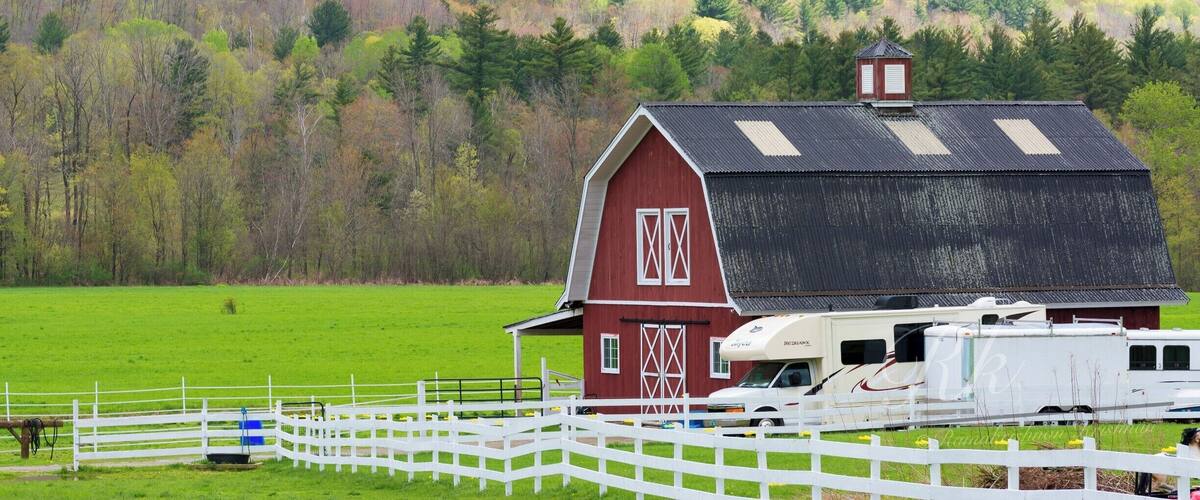 A red barn with a white fence in a green field with a white RV parked in front.