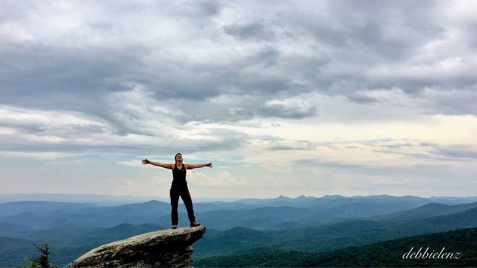 A person standing on a rock with arms outstretched, overlooking a mountainous landscape.
