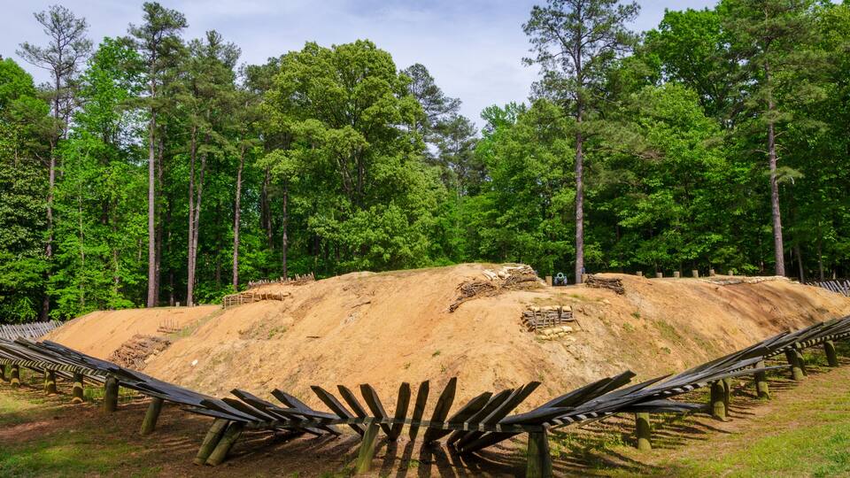 A large mound of earth with a wooden fence in the foreground, surrounded by a forest of tall trees.