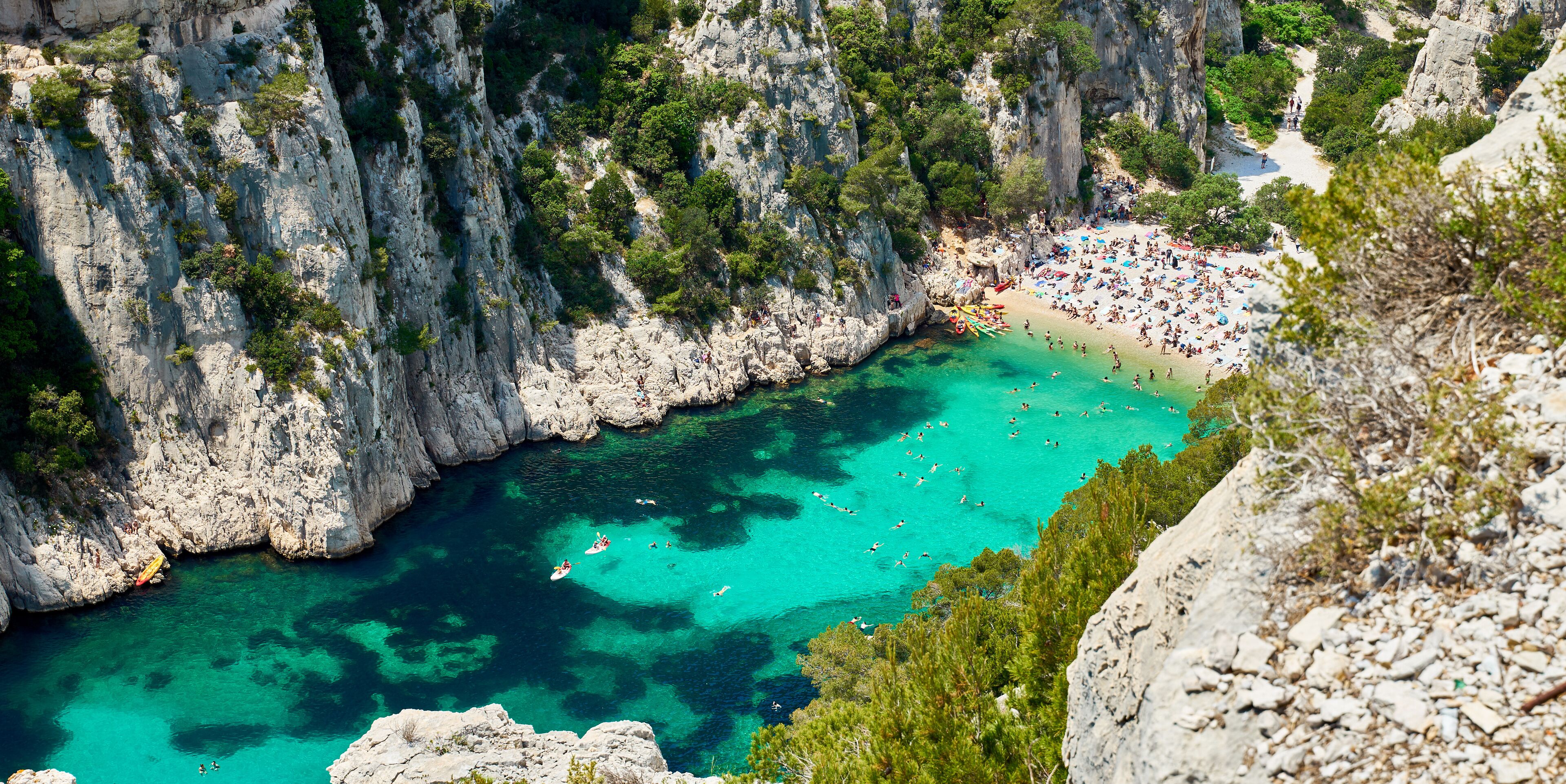 A coastal cliff with a turquoise bay, rocky cliffs, and a beach with people.