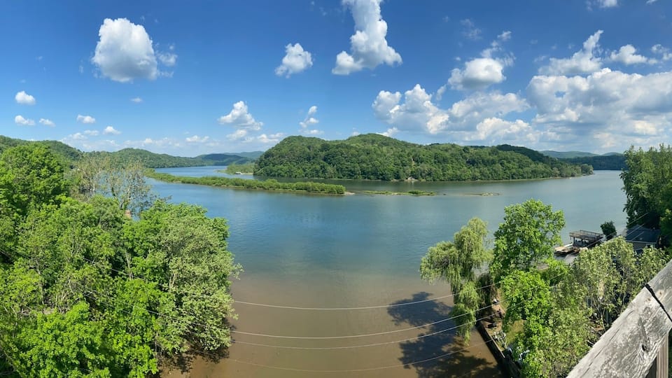 A wooden bridge over a calm river, surrounded by lush greenery and hills.