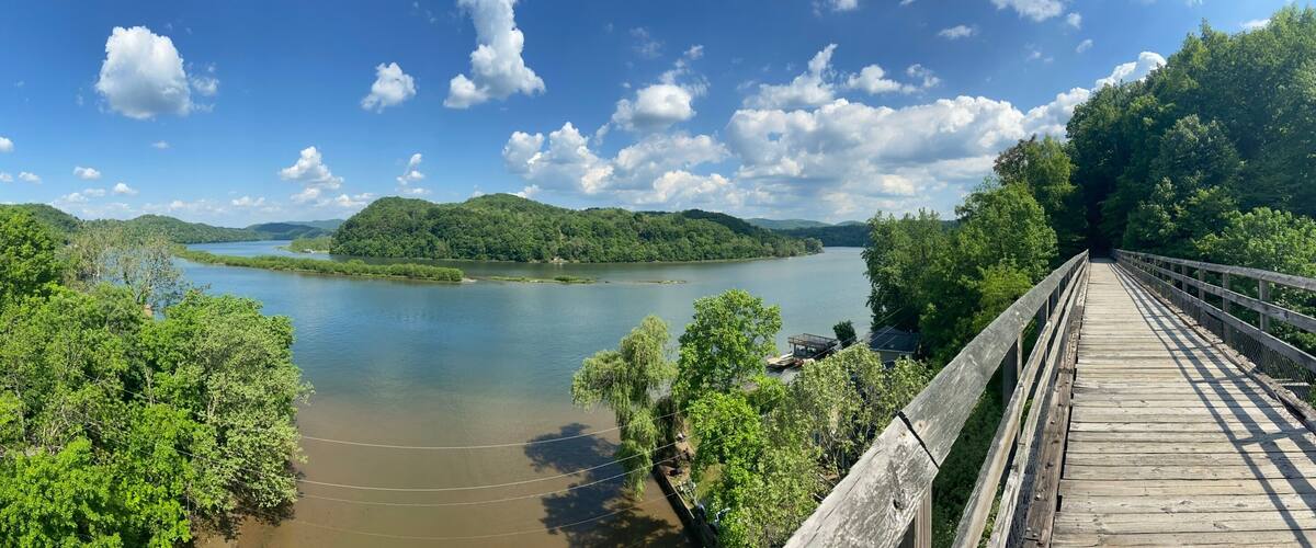 A wooden bridge over a calm river, surrounded by lush greenery and hills.