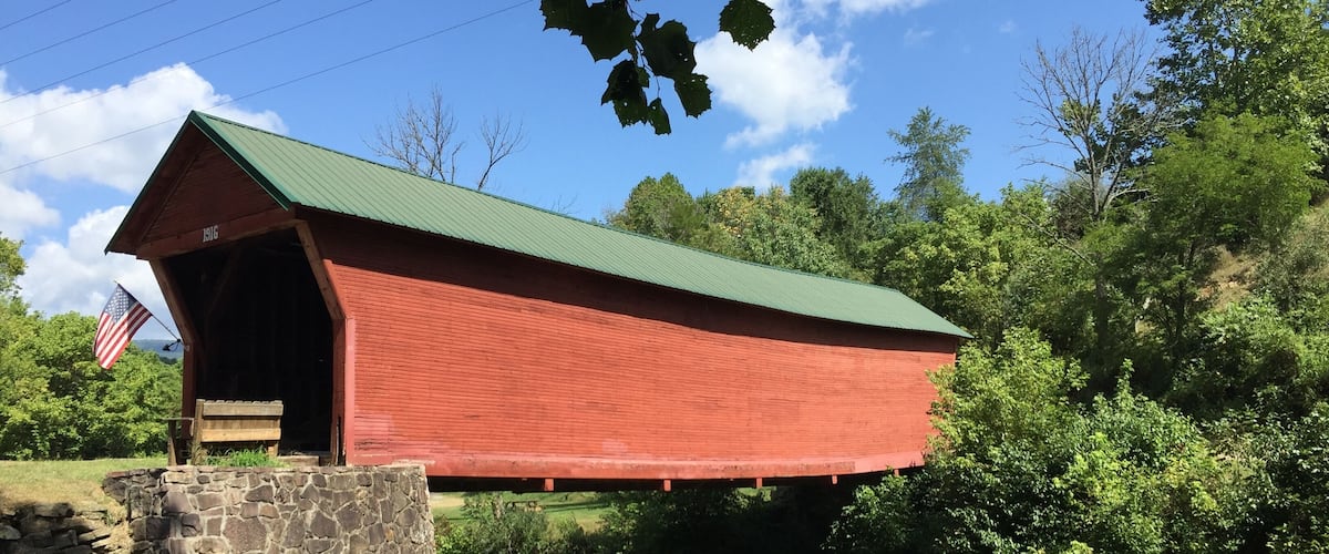 A red covered bridge with an American flag, a stone wall, and a river.