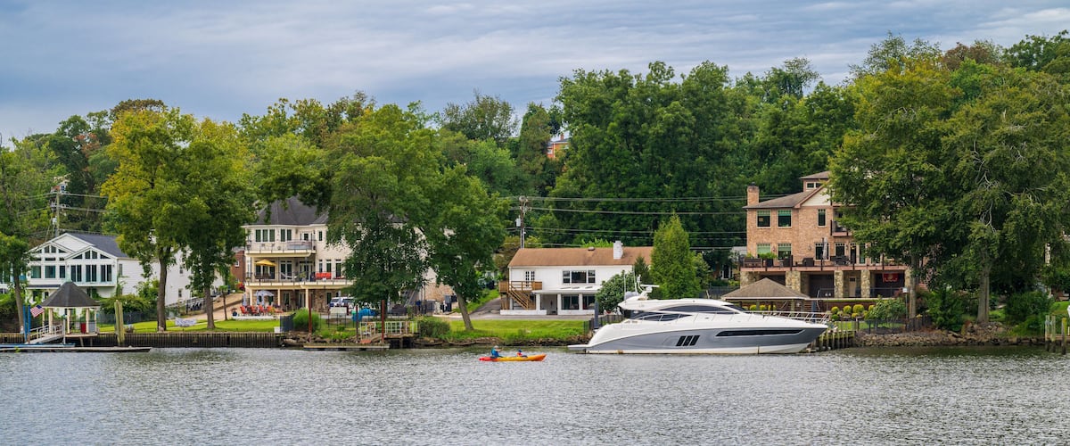 Une promenade en bord de mer avec un yacht amarré près d’une rangée de maisons, un kayak sur l’eau et un kiosque sur le quai.