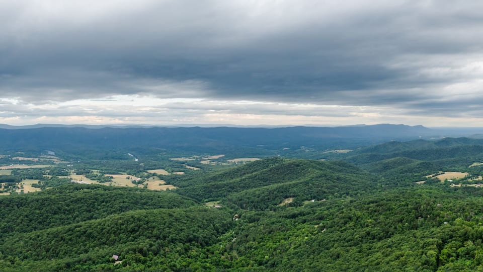 Aerial view of rolling green hills meeting the horizon under a stormy sky, with fields of gold and lush forests creating a rich tapestry, Rileyville, Virginia, USA.