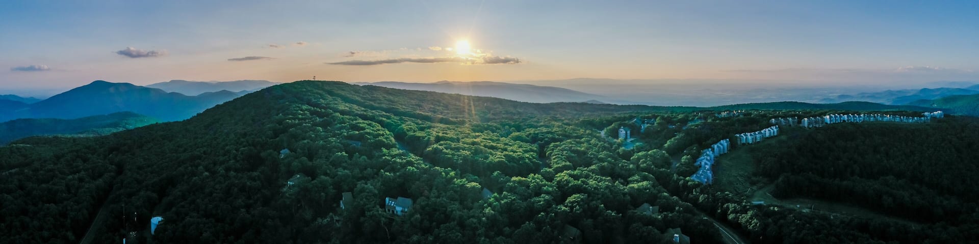 Aerial view of a sun-kissed mountain ridge covered in lush green trees, with a distant row of houses nestled among the foliage, Rileyville, Virginia, USA.